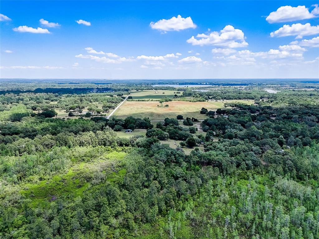 An aerial view shows a large, open field surrounded by dense forest and distant water — 7119 STATE ROAD 33, CLERMONT, FL