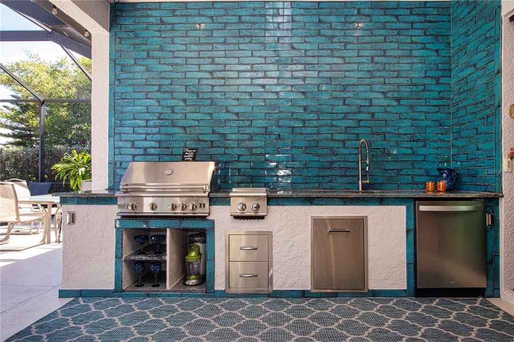 An outdoor kitchen features a teal brick backsplash, stainless steel appliances, and a patterned rug — 1929 24TH STREET CIRCLE W, PALMETTO, FL