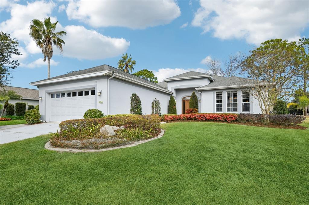 A light gray house exterior features a wooden front door, arched entryway, and landscaped garden — 8013 PINE HOLLOW DRIVE, MOUNT DORA, FL