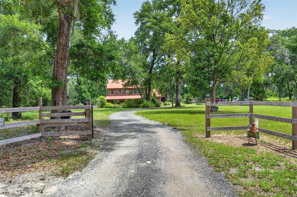 A gravel driveway leads through a wooden fence toward a two-story house among trees — 20621 SHADY OAK LANE, EUSTIS, FL