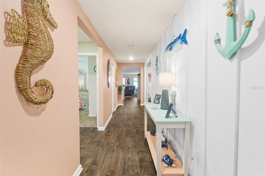 A hallway with wood-look flooring, peach walls, and coastal-themed decor leads to an open living area — 1 RICHFIELD PLACE, PALM COAST, FL