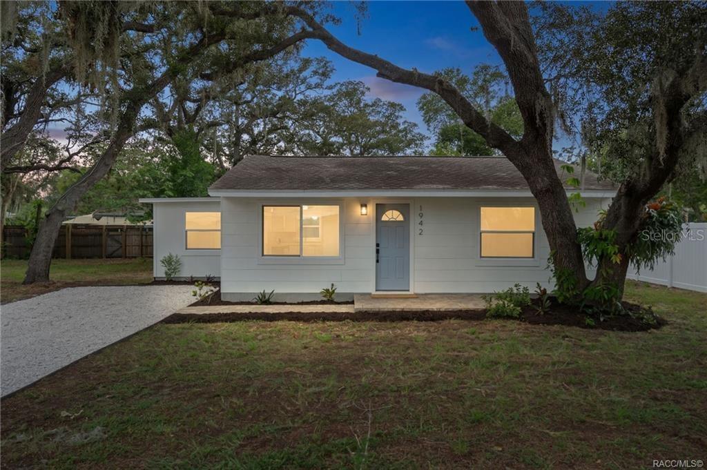 A single-story white house with a gray door and gravel driveway sits under large trees — 1942 SPRINGTIME AVENUE, CLEARWATER, FL