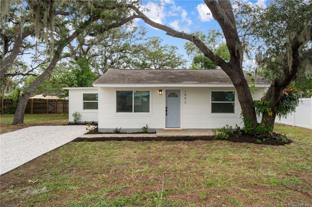 A white single-story house with a gray door and a gravel driveway under large trees — 1942 SPRINGTIME AVENUE, CLEARWATER, FL