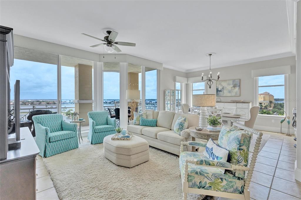 A bright living room features a beige sofa, patterned armchairs, and floor-to-ceiling windows with water views — 140 RIVIERA DUNES WAY UNIT 503, PALMETTO, FL