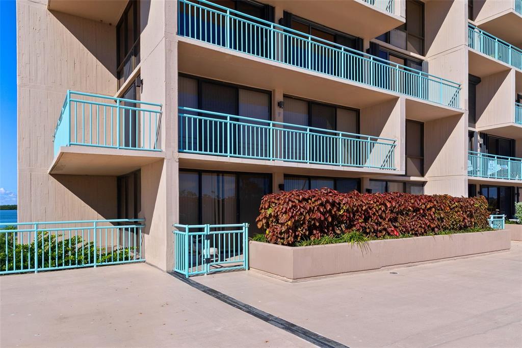A multi-story building exterior features light-colored walls, teal railings, and a ground-level patio area — 1 KEY CAPRI UNIT 102E, TREASURE ISLAND, FL