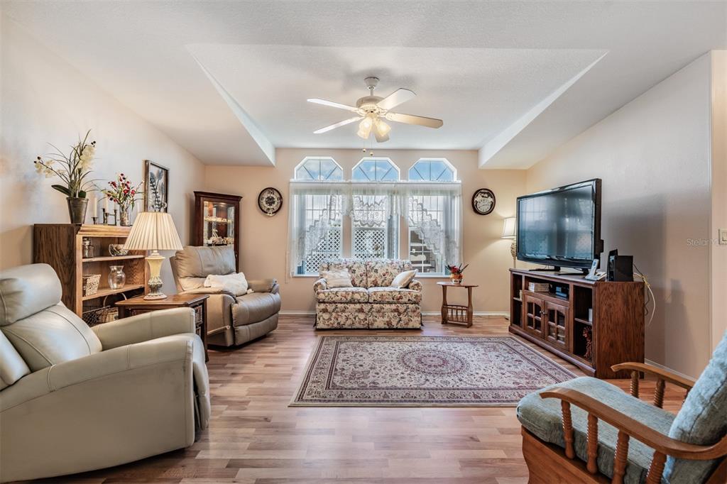 A living room features a tray ceiling, wood-look flooring, a floral loveseat, and a television — 37601 NEUKOM AVENUE, ZEPHYRHILLS, FL