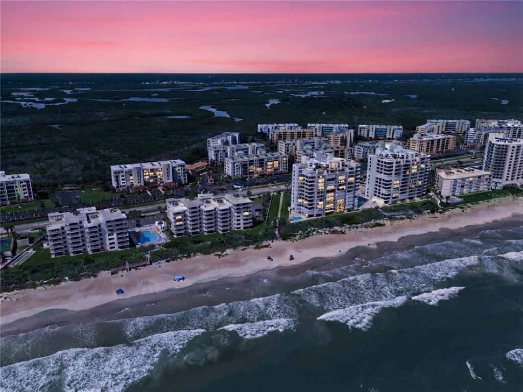 An aerial view shows several multi-story apartment buildings situated along a sandy beach at sunset — 5300 S ATLANTIC AVENUE UNIT 13-604, NEW SMYRNA BEACH, FL