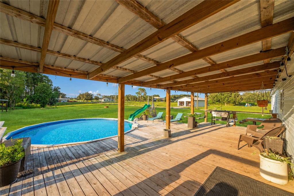 A wooden deck with a pool, slide, and covered patio area overlooking a grassy field — 3858 E KNIGHTS GRIFFIN ROAD, PLANT CITY, FL