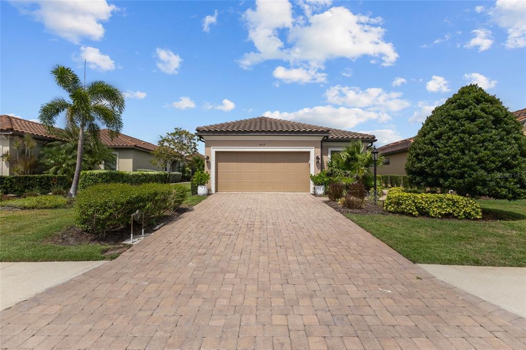 A single-story house with a tan garage, paver driveway, and landscaped yard under a blue sky — 5019 SAVONA RUN, BRADENTON, FL