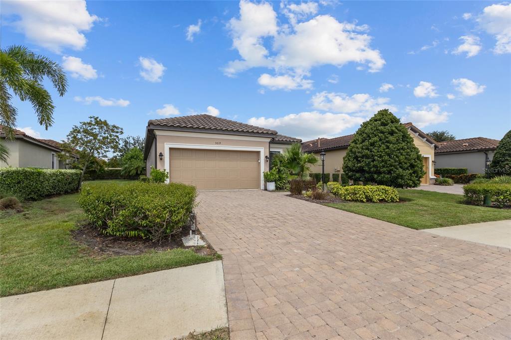 A single-story house with a tan garage, paver driveway, and manicured landscaping under a blue sky — 5019 SAVONA RUN, BRADENTON, FL