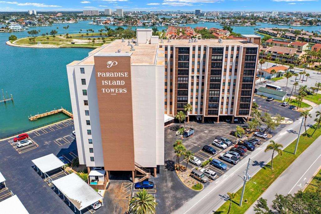 An aerial view shows a multi-story apartment building with a parking lot and waterfront access — 10355 PARADISE BOULEVARD UNIT 512, TREASURE ISLAND, FL
