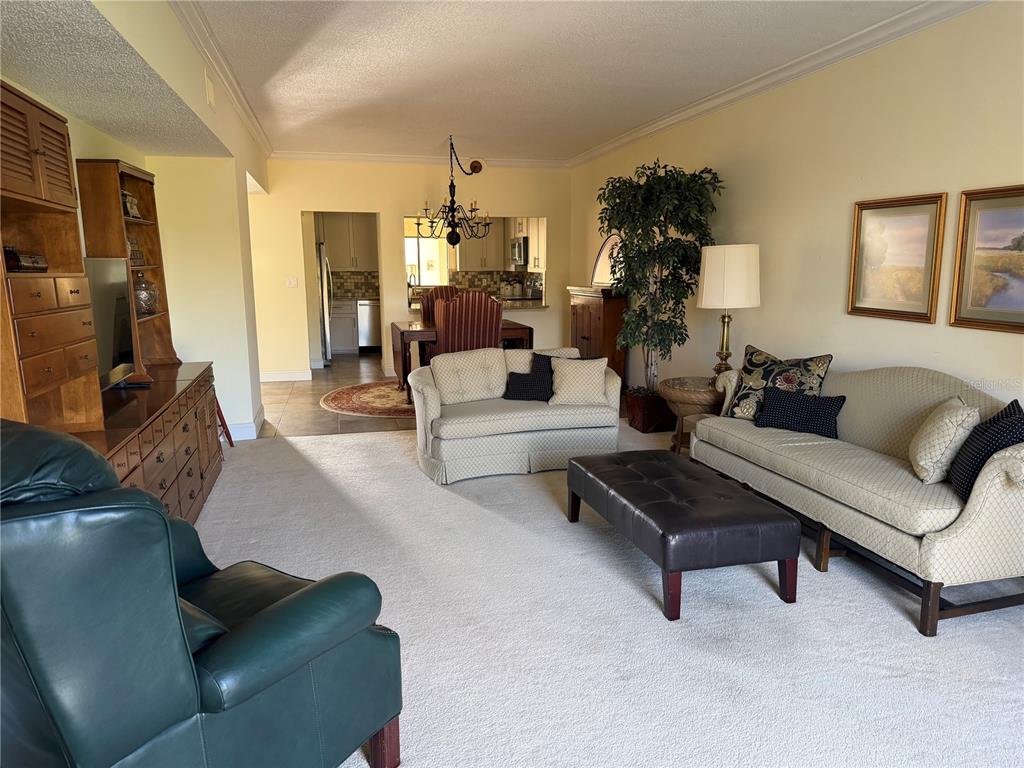 A living room with beige carpet, a leather ottoman, and a view into the dining area — 50 COE ROAD UNIT 322, BELLEAIR, FL