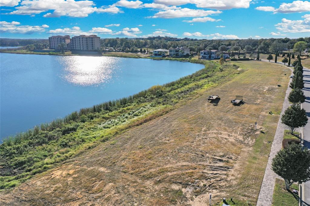 An aerial view shows a vacant grassy lot bordering a lake with distant residential buildings — 16626 BOLSENA DRIVE, BELLA COLLINA, FL