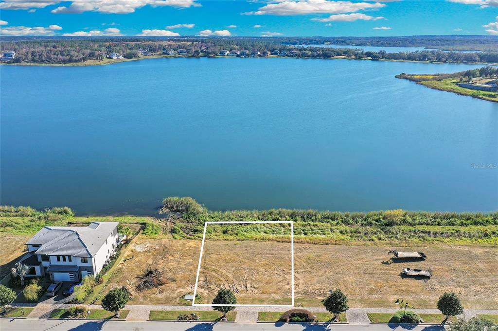 An aerial view shows a vacant rectangular lot situated along a lakefront with a neighboring house — 16626 BOLSENA DRIVE, BELLA COLLINA, FL