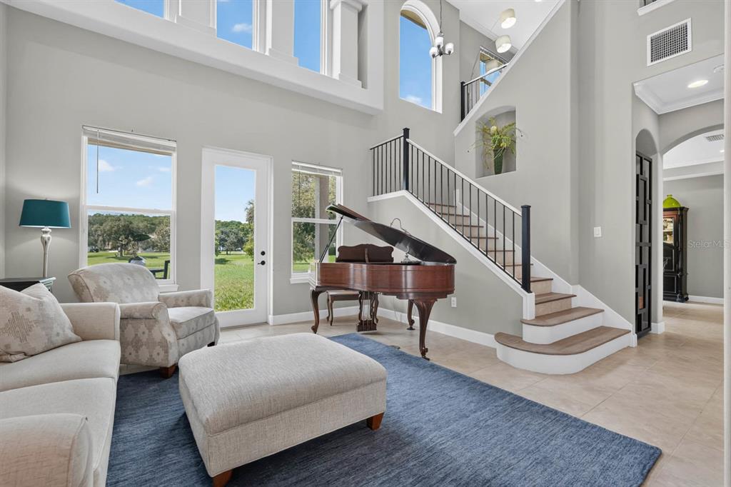 A living room features a grand piano, a staircase, and large windows overlooking a grassy yard — 55 NEW WATER OAK DRIVE, PALM COAST, FL