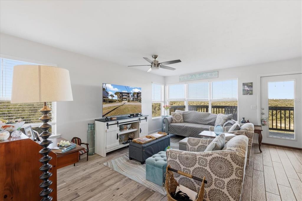 A bright living room features light wood-look tile flooring, a ceiling fan, and large windows — 11 OLD MILL DRIVE UNIT 9C, CEDAR KEY, FL