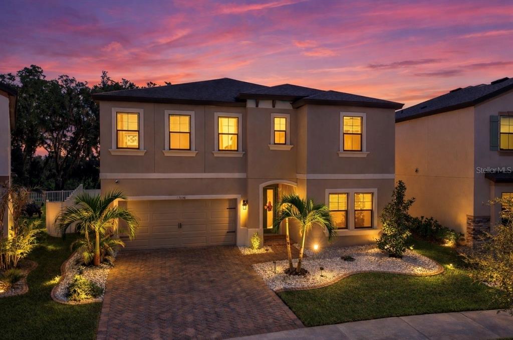 A two-story suburban house with a brick driveway, palm trees, and illuminated windows at sunset — 13036 PENNYBROOK DRIVE, RIVERVIEW, FL