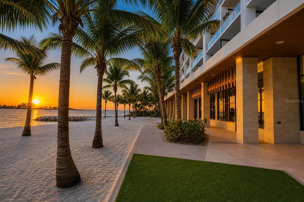 A modern building exterior features a sandy beach, palm trees, and a sunset view — 691 S GULFVIEW BOULEVARD UNIT 711, CLEARWATER, FL
