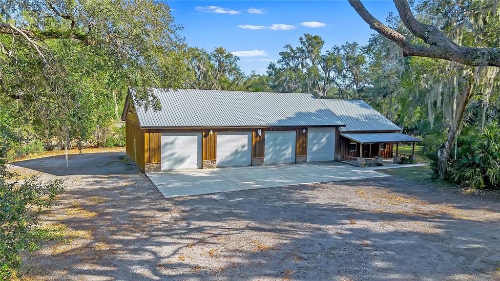 A large metal-roofed workshop with four garage doors sits on a gravel lot among trees — 27737 COUNTY ROAD 44A, EUSTIS, FL