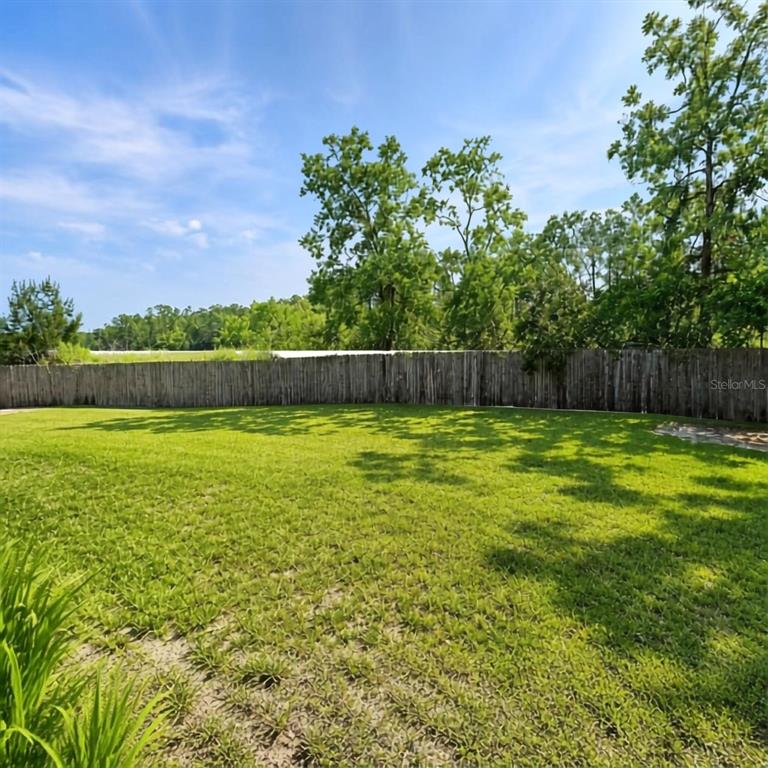 A grassy backyard features a wooden fence and mature trees under a blue sky — 1004 YALE DRIVE, DELTONA, FL