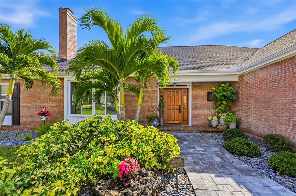A brick house exterior features a paved walkway, a wooden front door, and palm trees — 1307 RIVERVIEW CIRCLE, BRADENTON, FL