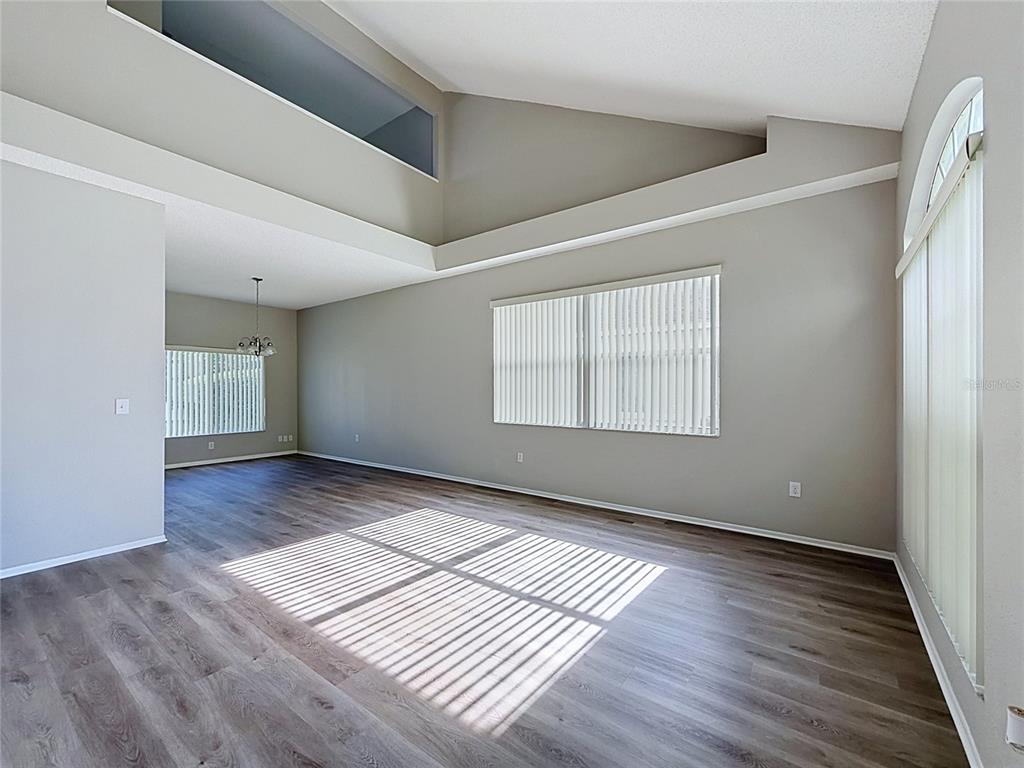 An empty living room features vaulted ceilings, gray wood-look flooring, and large windows with blinds — 27402 CORAL SPRINGS DRIVE, WESLEY CHAPEL, FL