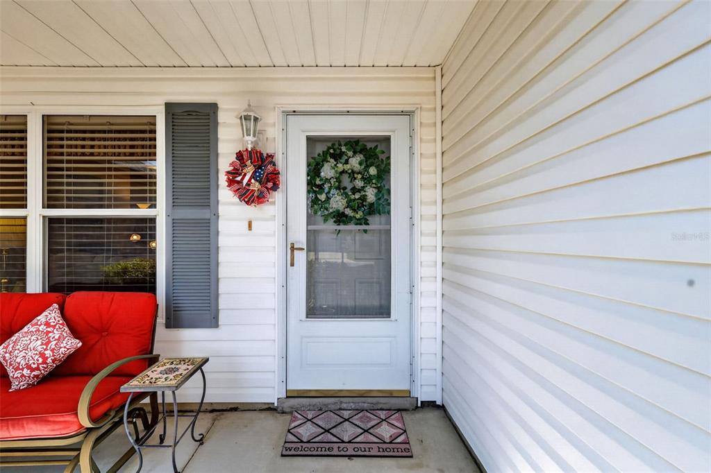 A front porch features white vinyl siding, a glass storm door, and a red patio chair — 1276 NORTHBROOK PLACE, THE VILLAGES, FL