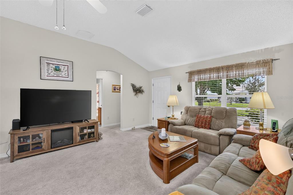 A living room features a large television, a wooden media console, and beige carpeted flooring — 1276 NORTHBROOK PLACE, THE VILLAGES, FL
