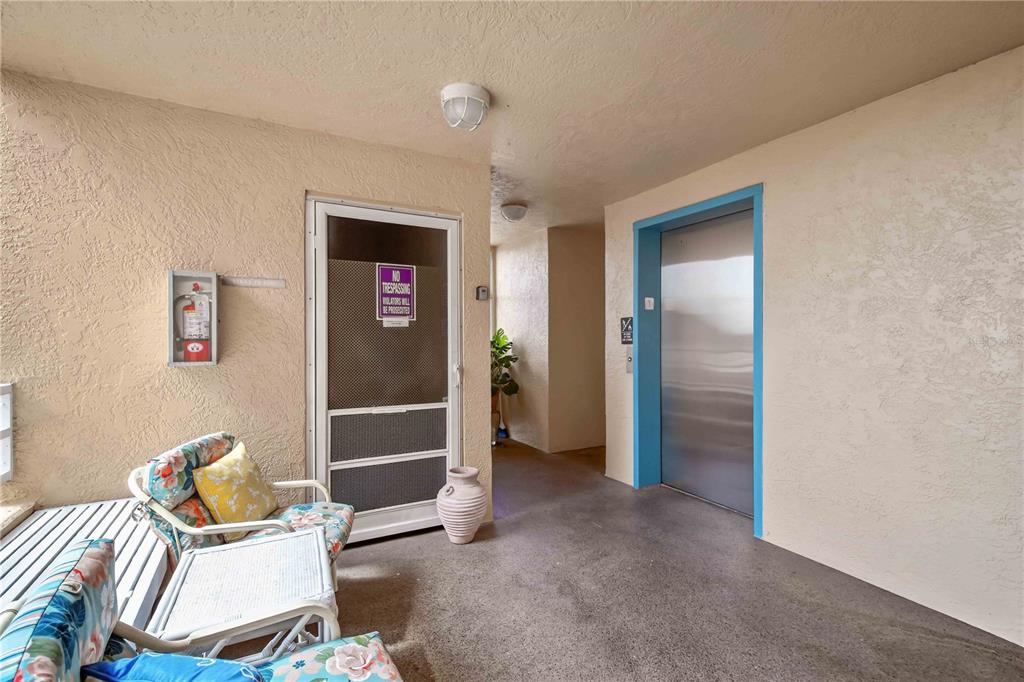 An outdoor hallway features a metal elevator, a screen door with a sign, and patio furniture — 4550 PINEBROOK CIRCLE UNIT 605, BRADENTON, FL