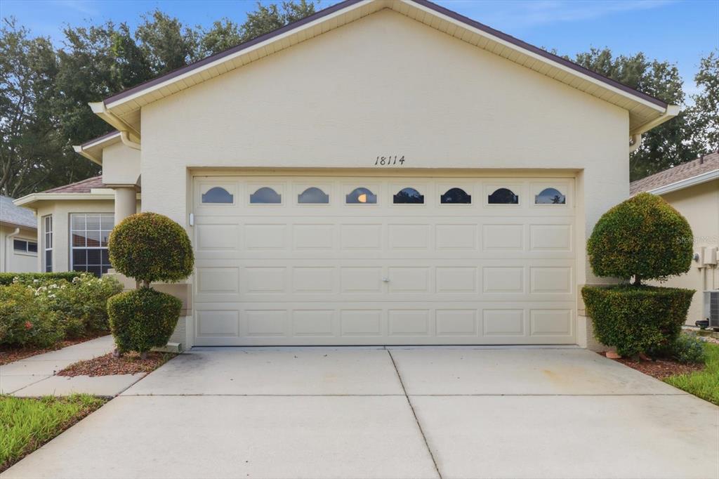 A beige house exterior features a two-car garage with arched windows and manicured topiary bushes — 18114 BRELAND DRIVE, HUDSON, FL