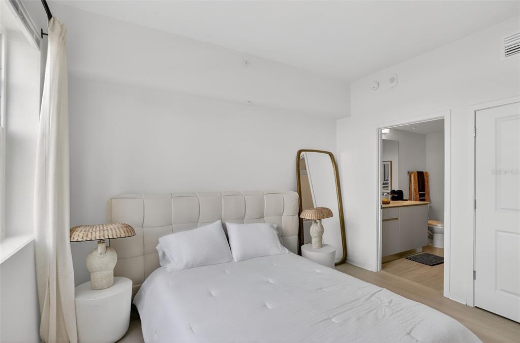 A bedroom features a tufted headboard, two bedside lamps, a floor mirror, and an attached bathroom — 5579 MILLENIA PARK BOULEVARD UNIT 402, ORLANDO, FL