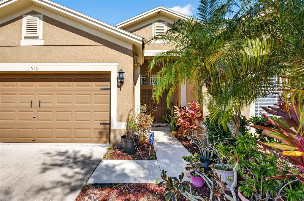 A tan house exterior features a two-car garage, a front walkway, and various potted plants — 11013 ROCKLEDGE VIEW DRIVE, RIVERVIEW, FL