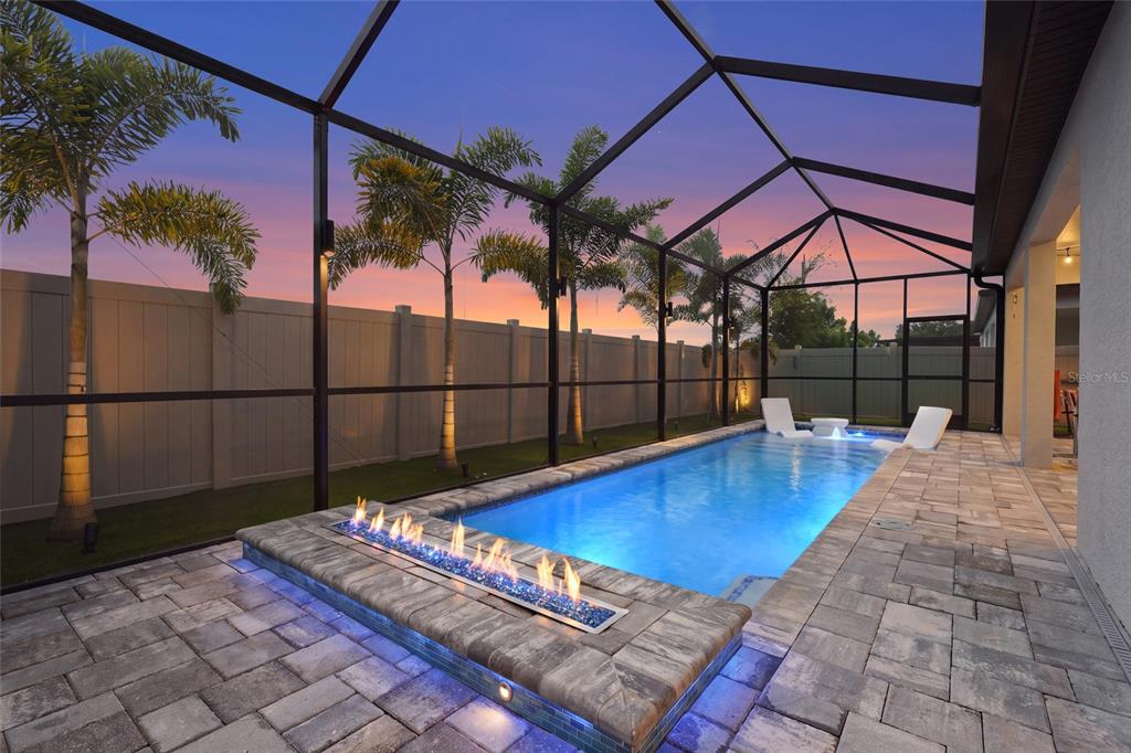 A screened-in pool area features a linear fire pit, stone pavers, and palm trees at sunset — 2930 61ST DRIVE E, ELLENTON, FL
