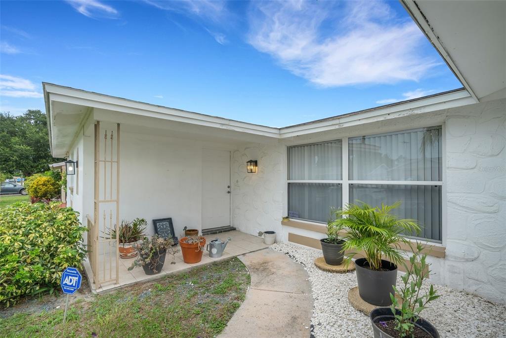 A white house exterior features a covered entryway, a walkway, and several potted plants — 7234 OAKSHIRE DRIVE, PORT RICHEY, FL