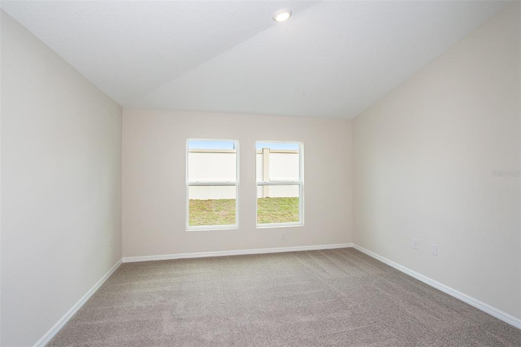 An empty bedroom features beige walls, gray carpeting, and two windows overlooking a grassy yard — 36691 THORNHEDGE DRIVE, ZEPHYRHILLS, FL