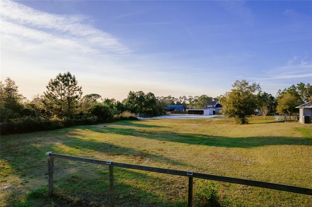 A grassy backyard features a wooden fence, scattered trees, and a distant outbuilding under sky — 7011 BRIDLE PATH, ST CLOUD, FL