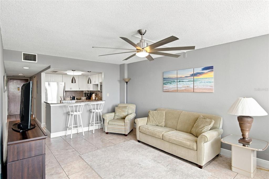 A living room with a sofa, armchair, ceiling fan, and an open kitchen with bar seating — 935 N HALIFAX AVENUE UNIT 604, DAYTONA BEACH, FL