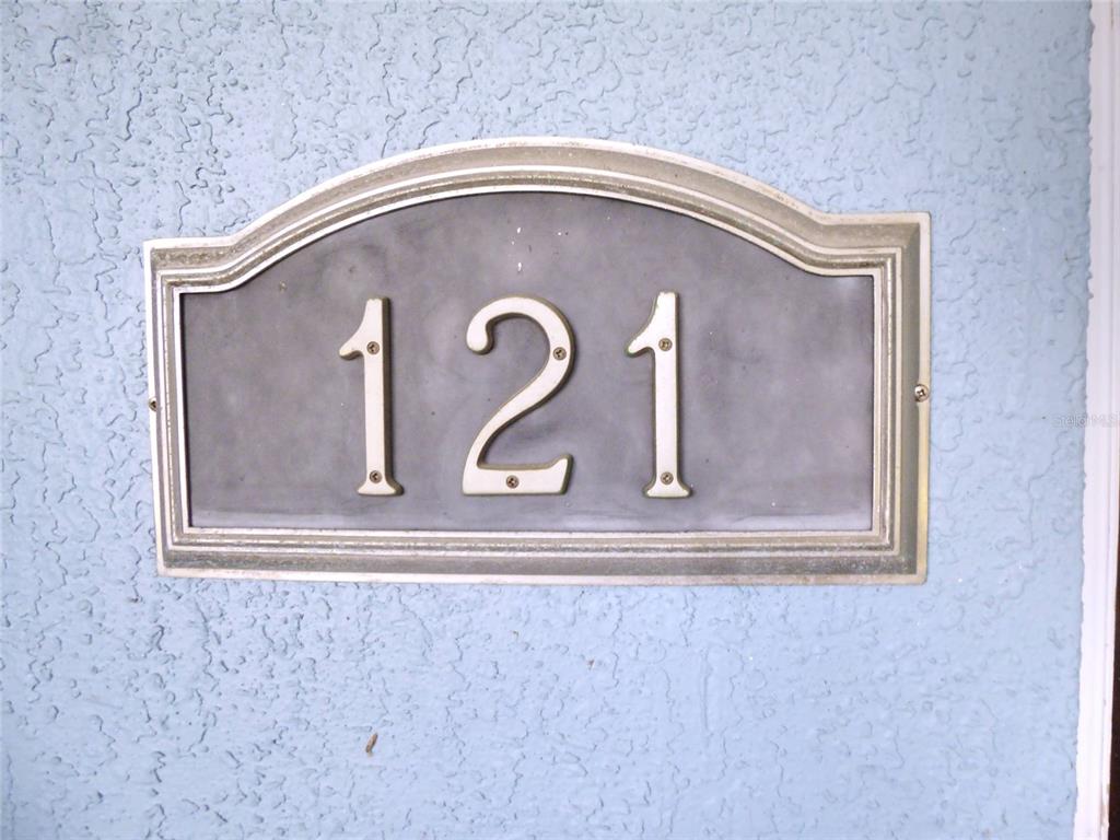 A house number plaque displaying the number 121 is mounted on a textured blue wall — 121 DEER ROAD, FROSTPROOF, FL