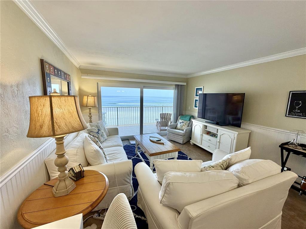 A living room features white furniture, a television, and a balcony overlooking the ocean — 4801 SAXON DR. UNIT A 202, NEW SMYRNA BEACH, FL