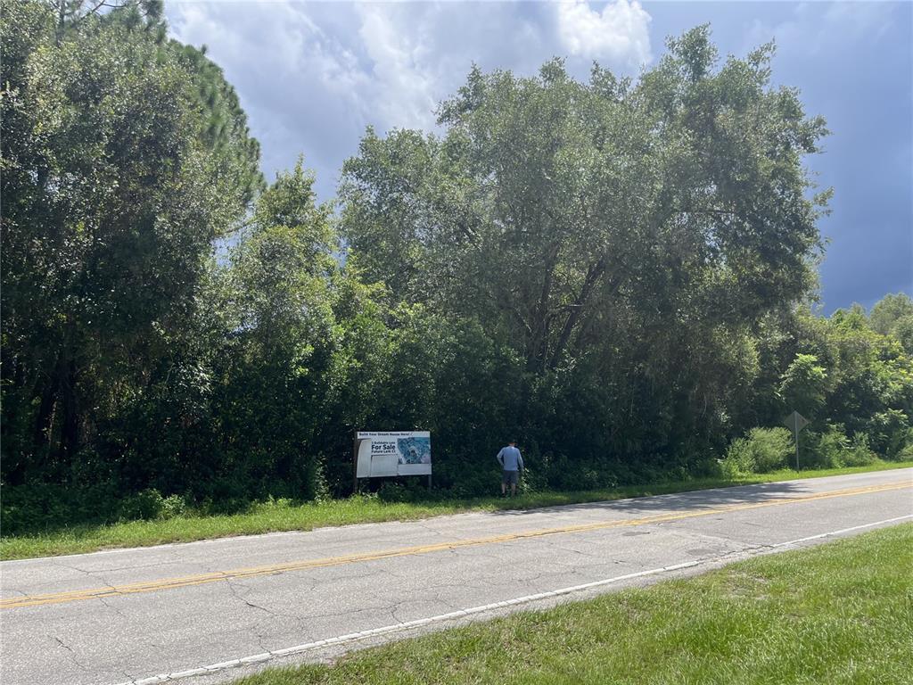 A person stands next to a for sale sign in front of a wooded lot — 847 LARK COURT, DELTONA, FL