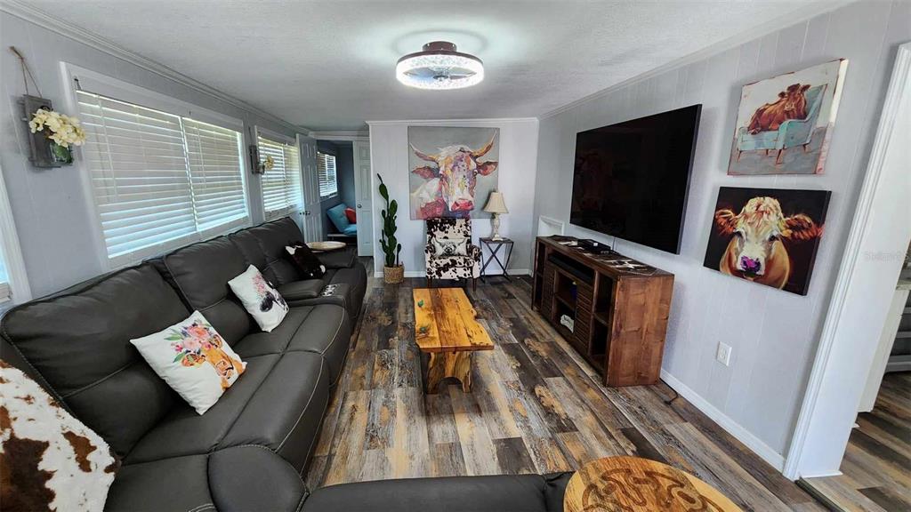 A living room features gray wood-look flooring, a dark leather sofa, and cow-themed wall art — 53 PALOMINO PATH, RIVER RANCH, FL