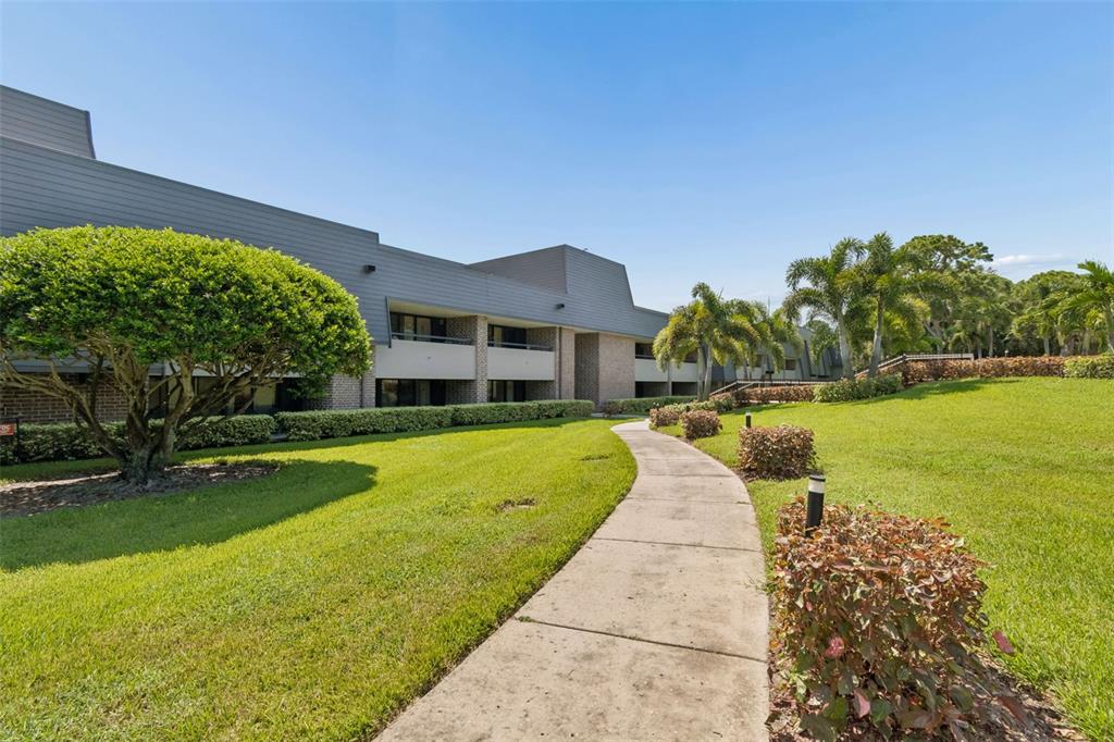 A concrete walkway leads past a manicured lawn and landscaping toward a multi-story apartment building — 36750 US HIGHWAY 19 N UNIT 04-125, PALM HARBOR, FL