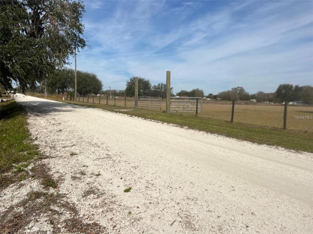 A gravel road runs alongside a fenced pasture with a metal gate under a blue sky — HART ROAD, WAUCHULA, FL