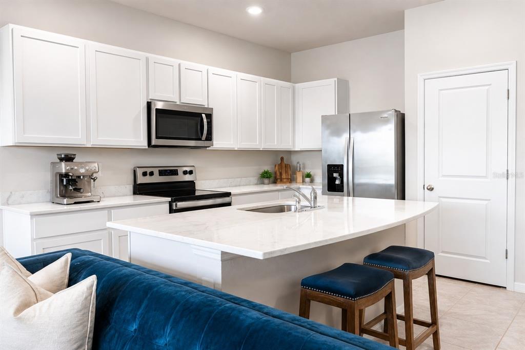 A modern kitchen features white cabinetry, stainless steel appliances, a marble island, and blue stools — 17632 CRESCENT MOON LOOP, BRADENTON, FL