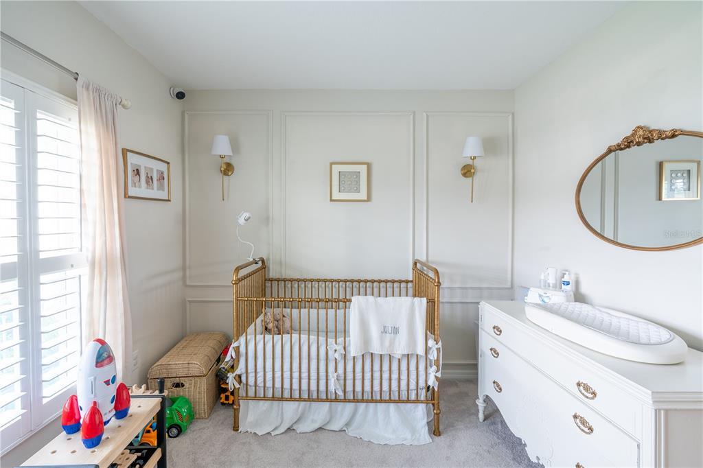 A nursery features a gold crib, white dresser, wall molding, and a window with shutters — 16261 YELLOWEYED DRIVE, CLERMONT, FL