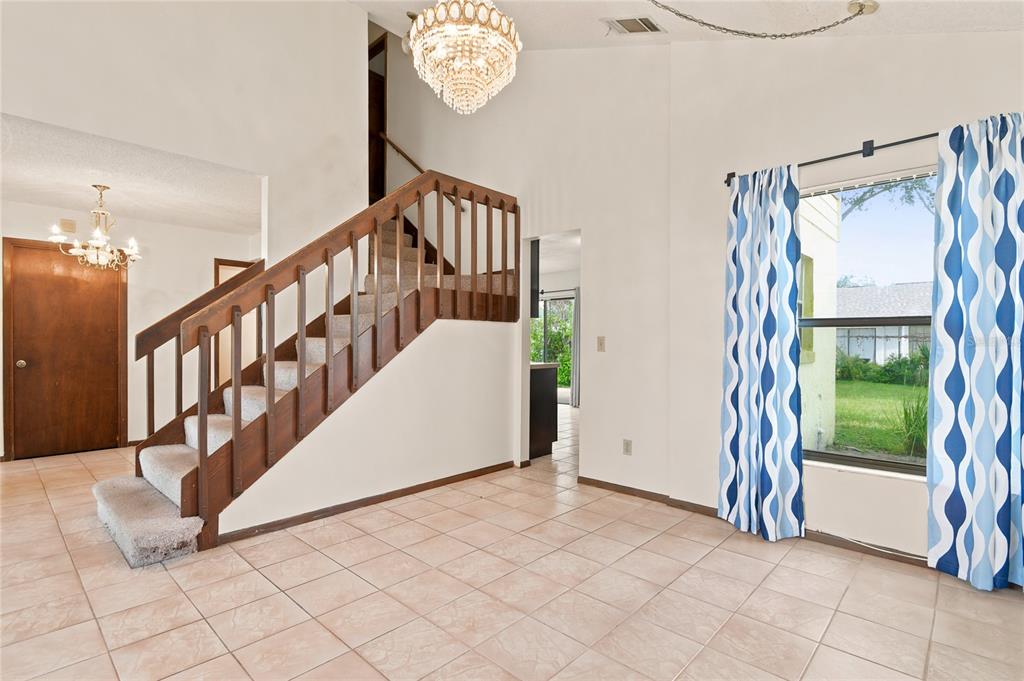 A tiled entryway features a wooden staircase, a crystal chandelier, and a window with curtains — 1469 MONA DRIVE, KISSIMMEE, FL