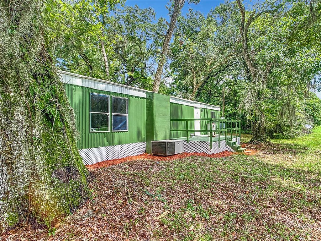 A green mobile home with a wooden deck sits in a wooded area — 13026 SW WILLISTON ROAD, MICANOPY, FL