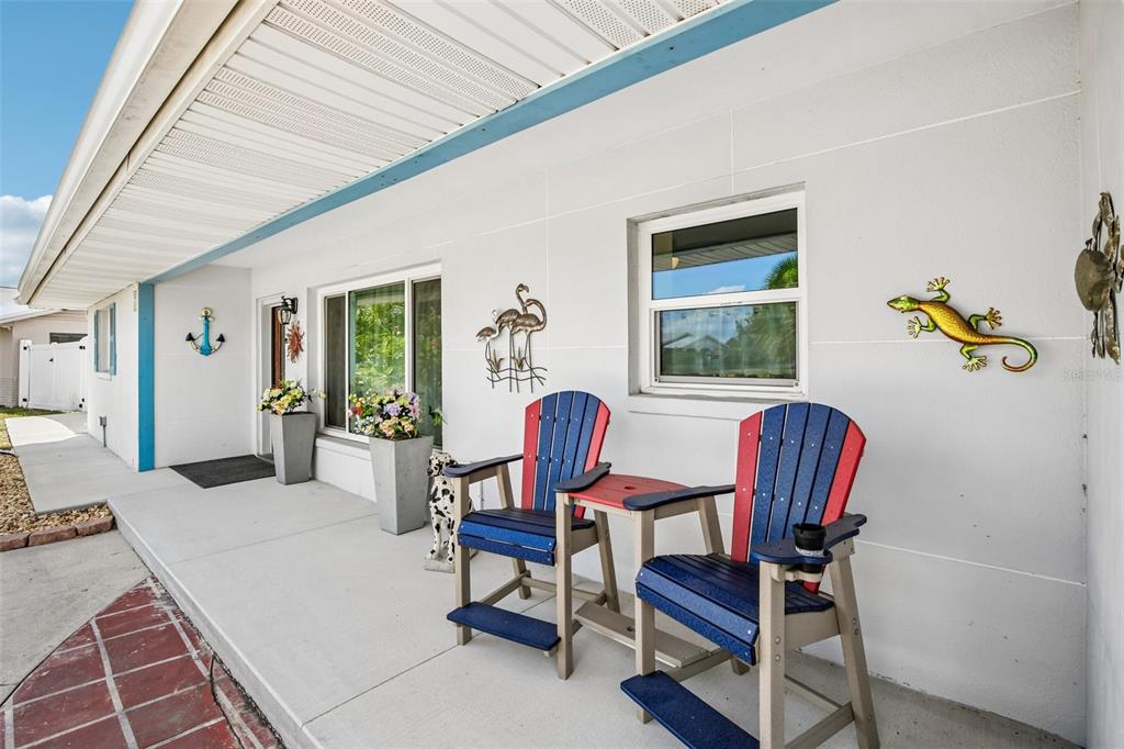 A covered concrete porch features two blue and red chairs, potted flowers, and wall decorations — 806 BLUE HERON BOULEVARD, RUSKIN, FL