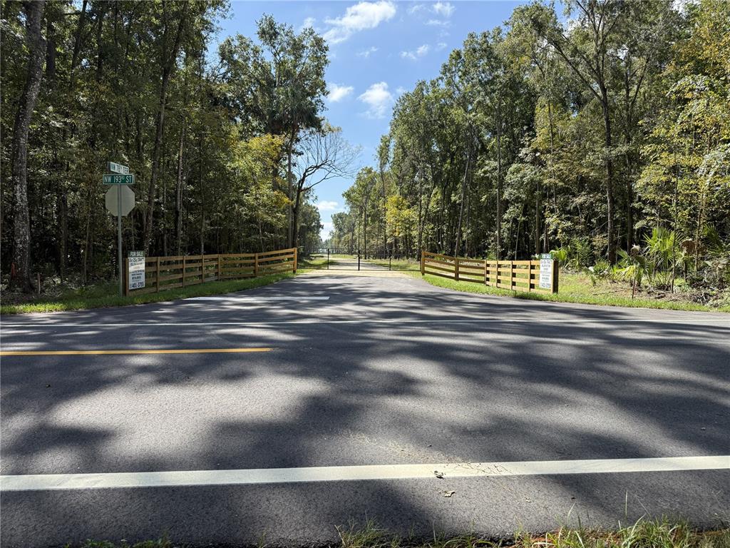 A paved road leads to a gated entrance flanked by wooden fences and dense trees — DR #7 (10ac) NW 193RD ST, MICANOPY, FL