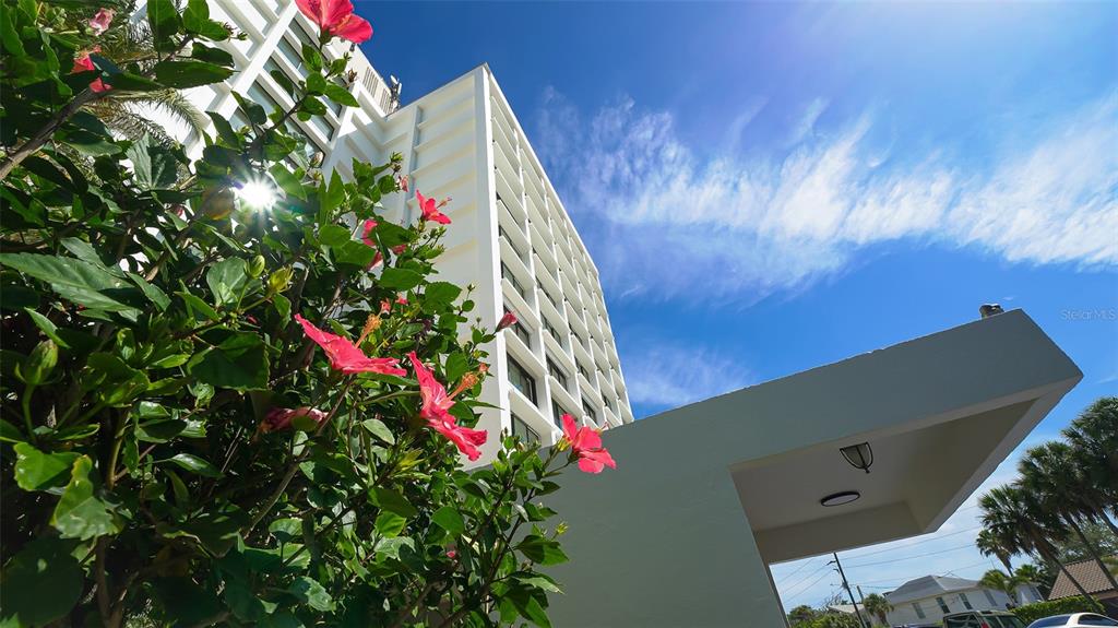 A low-angle view of a white high-rise building exterior with hibiscus flowers in the foreground — 4822 OCEAN BOULEVARD UNIT 2F, SARASOTA, FL